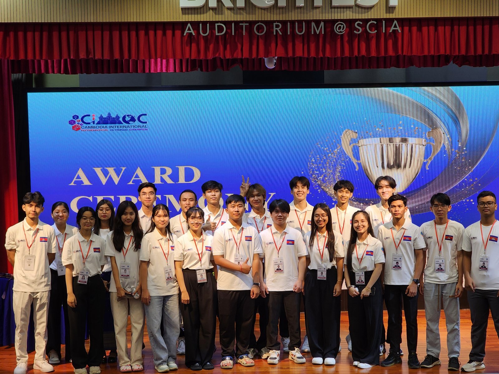 About twenty CIMOC volunteers in white polo shirts with Cambodian-flag badges and red lanyards, standing together on the SCIA auditorium stage in front of the Award Ceremony screen.