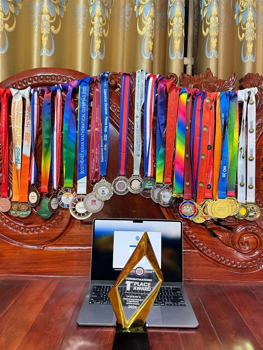 A display of mathematics olympiad medals hanging in rows — colorful ribbons from SEAMO 2023, Copernicus Olympiad 2022, and other competitions — with a golden first-place Angkor Mathematics Competition 2024 trophy standing in front of a laptop.