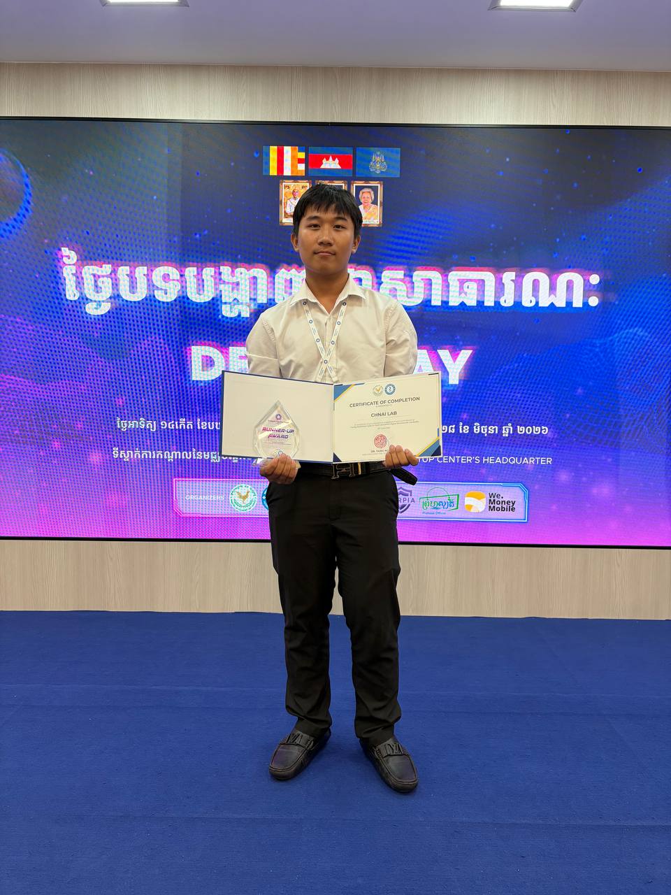 Chamroeun Hongleng standing on the Demo Day stage holding an open folder with the CHNAI LAB certificate of completion and the Runner-Up Award trophy.
