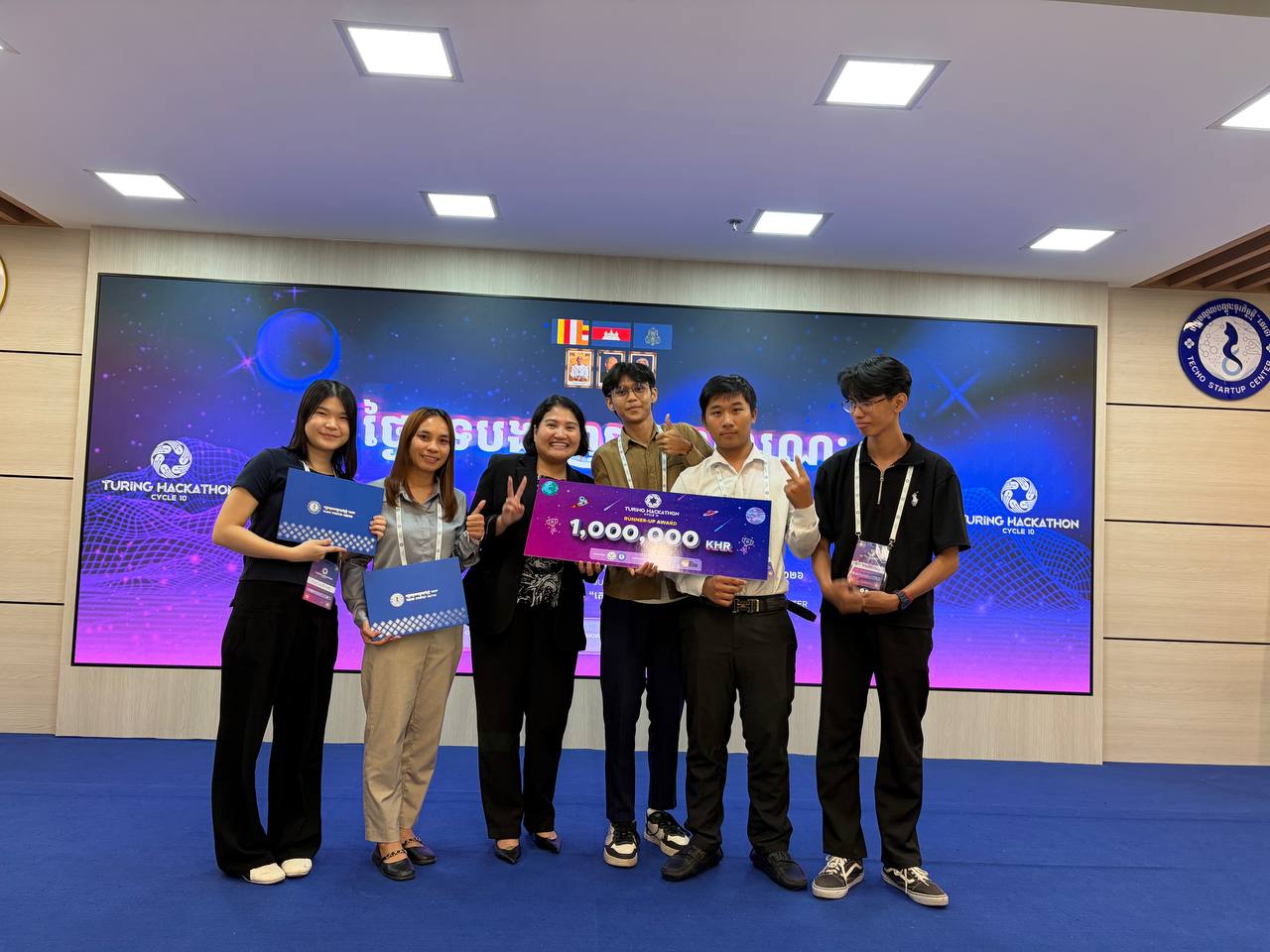 Six people standing on the Techo Startup Center stage in front of a Turing Hackathon Cycle 10 screen, holding certificate folders and an oversized 1,000,000 KHR Runner-Up Award cheque.